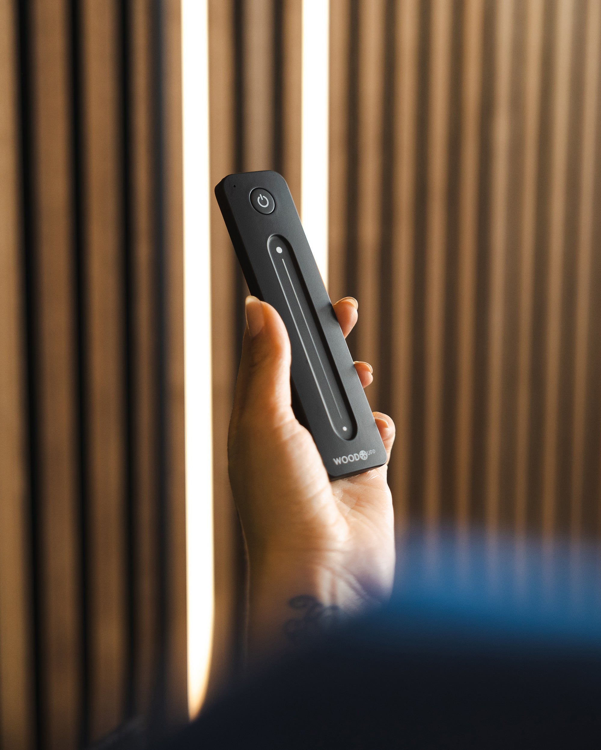 A hand holds a black remote control with a power button, positioned near vertical Akupanel acoustic wood slats.